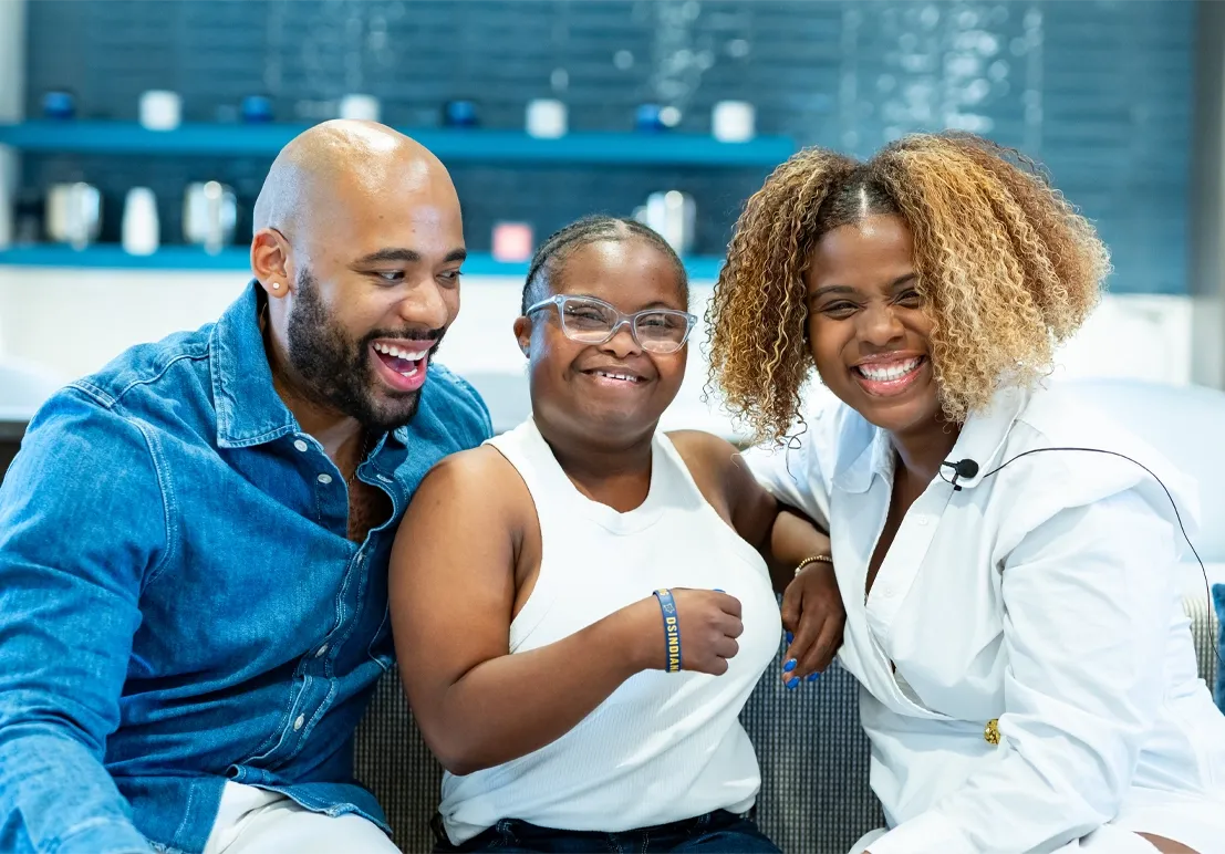 Man, young woman with Down syndrome, and woman laughing together on a couch, representing the Indiana Disability Alliance community