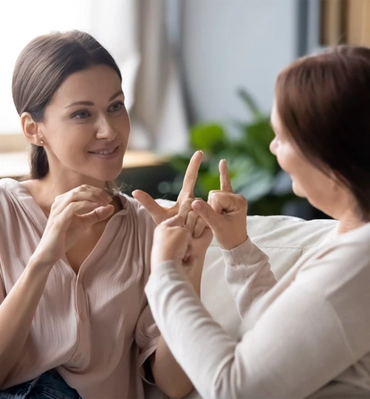Two women communicating using American Sign Language in a warm living room setting, reflecting accessibility and community connection